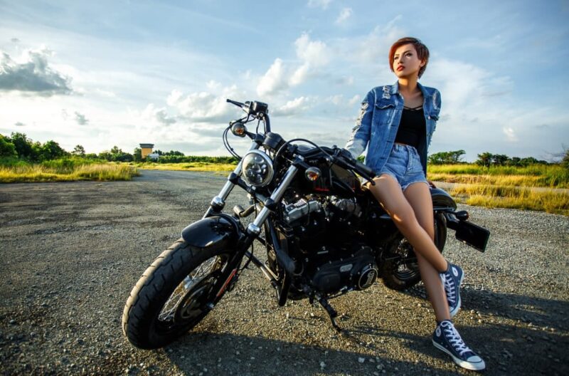 A woman sitting on a black cruiser motorcycle in a denim jacket and shorts, with a field in the background