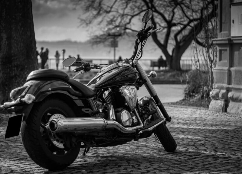 A black and white image of a classic motorcycle parked on a cobblestone street by the riverside