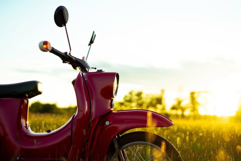 A vintage red scooter parked in a field with the sun setting in the background